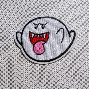 May include: Embroidered patch featuring a cartoon ghost with a white body, black outline, and a red mouth with a pink tongue. The ghost has two small, black eyes and two small arms. The patch is on a patterned background.