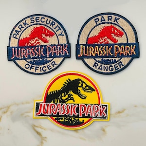 May include: Three embroidered patches featuring the Jurassic Park logo. The first patch is blue and white with the text "Park Security Officer" and a red dinosaur. The second patch is blue and white with the text "Park Ranger" and a red dinosaur. The third patch is yellow with a black dinosaur skeleton and the text "Jurassic Park".