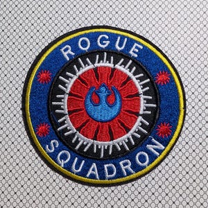 May include: Embroidered patch with the text "ROGUE SQUADRON" in white on a blue circular background. The patch features a red and blue emblem in the center, with yellow and red accents. The patch is likely for a fan of a science fiction franchise.