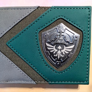 May include: A bi-fold wallet featuring a combination of grey and teal faux leather panels with a central olive green canvas section. A silver-toned metal emblem shaped like a shield with a bird and triangle design is attached to the front.