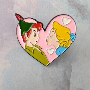 May include: A heart-shaped enamel pin featuring Peter Pan and Wendy from the Disney movie. Peter Pan is in a green outfit with a red feather, and Wendy is in a blue dress. The background is pink with white heart accents.