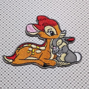 May include: Embroidered patch featuring Bambi and Thumper. Bambi is depicted in shades of orange, red, and yellow, with a pink nose and blue eyes. Thumper is gray and white. The patch is outlined in gray.