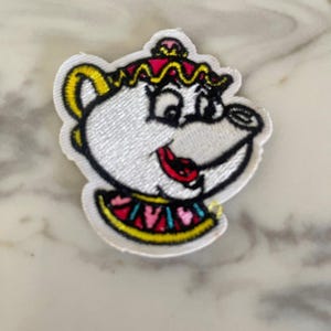 May include: White embroidered iron-on patch of a cartoon teapot with a pink and yellow crown, pink and blue collar, and a red tongue sticking out.