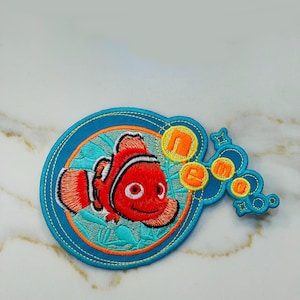 May include: An embroidered patch featuring a cartoon orange and white clownfish with a blue and green background. The fish is surrounded by bubbles that spell out "Nemo".