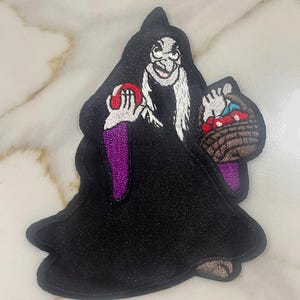 May include: An embroidered patch of a woman in a black robe holding a red apple in one hand and a basket of apples in the other. The woman has a white beard and a sinister smile.