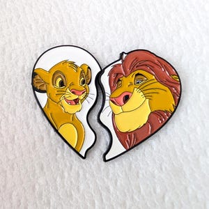 May include: Two enamel pins, each shaped like a half-heart, featuring Simba and Mufasa from The Lion King. Simba is depicted as a cub, and Mufasa as an adult lion. The pins have a white border and are set against a white background.