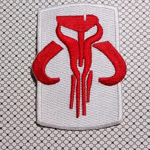 May include: Embroidered patch featuring a red Mandalorian symbol on a white background. The patch has a rectangular shape with rounded corners and a stitched border. The symbol is a stylized design with curved and vertical lines.