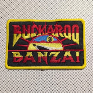May include: Embroidered patch with the words "BUCKAROO BANZAI" in red and yellow lettering. The patch features a stylized sun design with red rays and a blue, yellow, and red center, all on a black background with a yellow border.