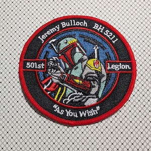 May include: Embroidered patch featuring a detailed illustration of a character in a helmet and armor, set against a blue background. The patch is circular, with text including "Jeremy Bulloch," "501st Legion," and the phrase "As You Wish."