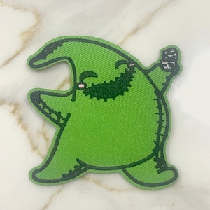 May include: Green felt patch of a cartoon character with a wide grin and a single arm raised. The character is wearing a green hat and has a stitched-on appearance.