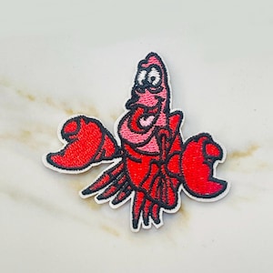 May include: Embroidered patch featuring a cartoon crab character in shades of red and pink with black outlining. The crab has large claws and a smiling expression. The patch is on a white background.