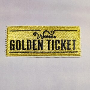 May include: Embroidered patch featuring a gold background with black text that reads "Wonka GOLDEN TICKET". The patch has a rectangular shape with a white border and a stitched edge. The patch is a collectible item.