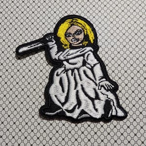 May include: Embroidered patch featuring a cartoon character with blonde hair, wearing a white dress, and holding a knife. The patch has a black border and is set against a patterned background.