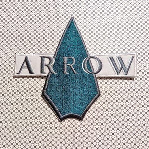 May include: Embroidered patch featuring the word "ARROW" in gray thread on a white rectangular background. The arrow is teal with a black and gray border. The background has a repeating pattern of small black squares.