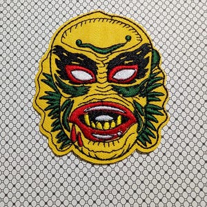 May include: A yellow embroidered patch featuring a monster face with red eyes, a red mouth with sharp teeth, and green accents. The patch has a black outline and is on a patterned background. The design is reminiscent of a classic horror movie creature.