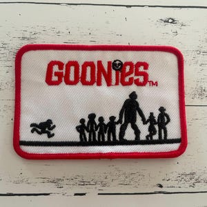 May include: A red, white, and black embroidered patch with the word "GOONIES" in red letters and a silhouette of a group of people running. The patch has a white background and a red border.