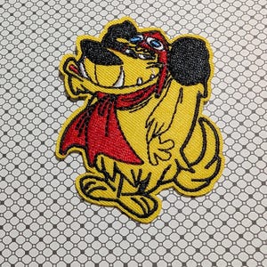 May include: Embroidered patch featuring a cartoon dog with yellow fur, black ears, and a red cape. The dog has a mischievous grin and blue eyes. The patch is outlined in black and is on a patterned background.