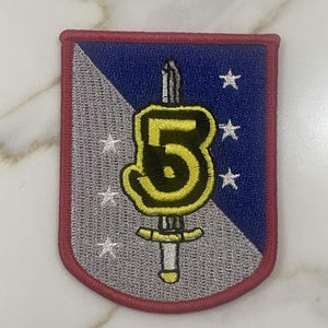 May include: A blue and gray embroidered patch with a red border. The patch features a yellow number "5" with a sword through it and five white stars.