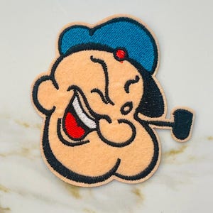 May include: An embroidered patch of Popeye the Sailor Man. He is wearing a blue sailor hat and has a red dot on his head. He is smiling and holding a pipe in his mouth.