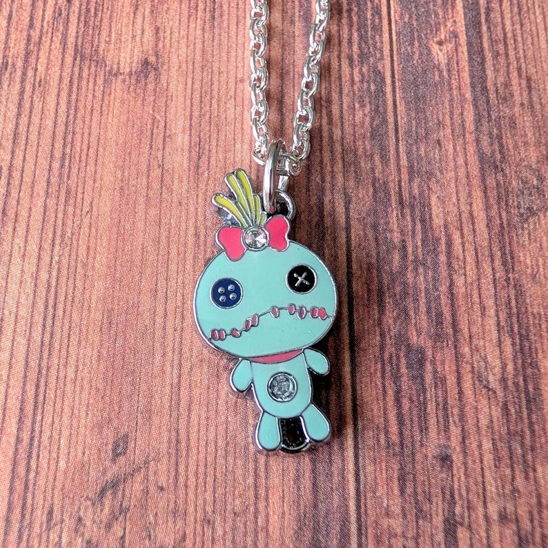 May include: A silver-toned necklace featuring a teal voodoo doll pendant. The pendant has a pink bow, button eyes, and a stitched mouth. The doll's design includes a small gem and a black cross. The necklace hangs on a silver chain.