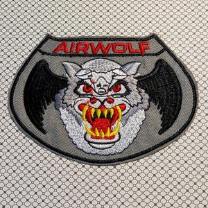 May include: Embroidered patch featuring a snarling wolf with red eyes and yellow fangs, set against a gray shield shape. The word "AIRWOLF" is arched across the top in red letters. Black wings extend from the wolf's head.
