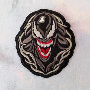May include: Embroidered patch featuring a stylized monster face with a wide, red-lined mouth and sharp teeth. The design includes gray and white accents on a black background, with a black border. The patch is oval-shaped.