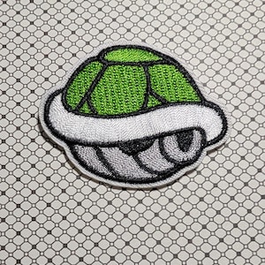 May include: Embroidered patch featuring a green turtle shell with a white border and gray accents. The shell has a textured appearance and is likely for clothing or accessories.