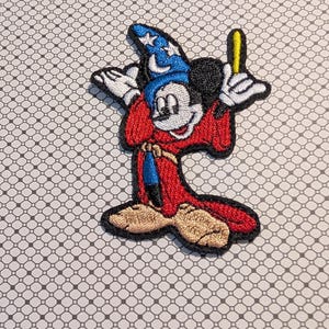 May include: Embroidered patch featuring Mickey Mouse as the Sorcerer's Apprentice. The patch shows Mickey in a red robe, blue wizard hat with stars, and holding a yellow wand. The patch has a black border.