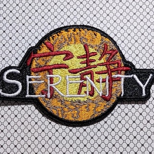May include: Embroidered patch with the word "SERENITY" in white and red Japanese characters. The patch has a black border and a yellow and orange sunburst design in the center. The background is a white and gray patterned fabric.