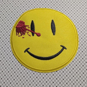 May include: Yellow embroidered patch featuring a smiley face with black eyes and a curved mouth. A red, stylized blood splatter design is over the right eye. The patch has a yellow border.