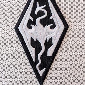 May include: A black and white embroidered patch in a diamond shape. The patch features a white dragon design on a black background. The design is detailed with intricate stitching, and the patch has a black border.