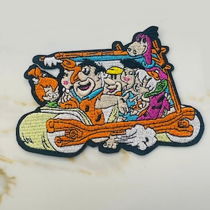 May include: Embroidered patch featuring the Flintstones in their iconic car. The patch showcases the characters in vibrant colors, including orange, blue, pink, and yellow, with a black border. The design is detailed and suitable for application on clothing or accessories.
