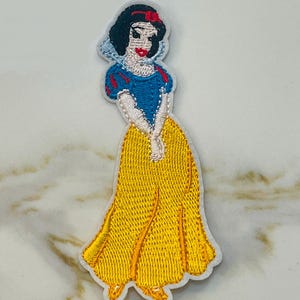 May include: An embroidered iron-on patch featuring Snow White from Disney's Snow White and the Seven Dwarfs. The patch is white with a yellow dress, blue bodice, and red bow.