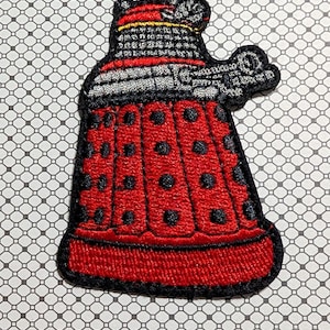 May include: Embroidered patch featuring a red Dalek design with black polka dots and silver and black details. The patch has a black border and is likely for application on clothing or accessories. The design is based on the iconic science fiction character.