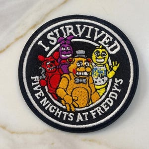 May include: A black and white embroidered patch with the text "I Survived Five Nights at Freddy's". The patch features four cartoon characters from the game, including Freddy Fazbear, Bonnie the Bunny, Chica the Chicken, and Foxy the Pirate Fox.