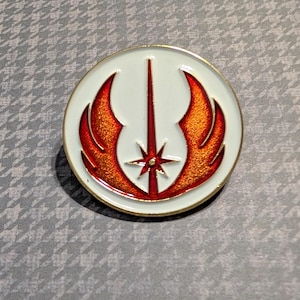 May include: A round, white and gold pin featuring a red and orange emblem with a central vertical line and a starburst at the bottom. The emblem is surrounded by a gold border. The pin is set against a gray patterned background.