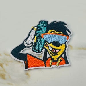 May include: Embroidered iron-on patch featuring a cartoon dog wearing sunglasses and a gold chain. The dog is holding a blue and green object in its right hand. The patch has a white background and is surrounded by a white border.