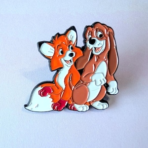 May include: A colorful enamel pin featuring Tod the fox and Copper the hound dog from the animated film, The Fox and the Hound. The pin has a white, orange, red, and brown color scheme. The fox is on the left, and the dog is on the right.