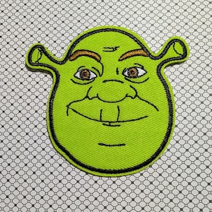 May include: A green embroidered patch featuring the face of Shrek, with brown eyes, a black outline, and a smiling expression. The patch has a textured appearance and is likely designed to be ironed or sewn onto fabric.