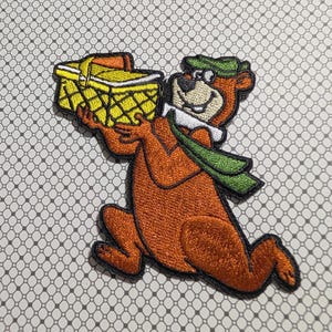 May include: Embroidered patch featuring a cartoon bear in brown, wearing a green hat and scarf, holding a yellow picnic basket with an orange handle. The patch has a black border and is on a patterned background.