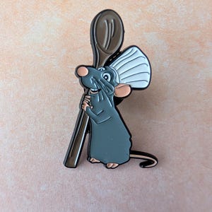 May include: A gray cartoon rat pin holding a brown wooden spoon. The rat has large ears and a long tail. The pin is set against a peach-colored background.