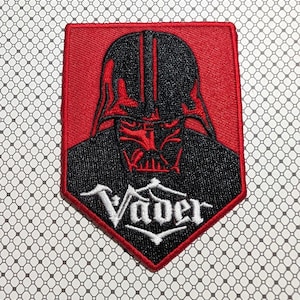 May include: Embroidered patch featuring Darth Vader's likeness in black and red. The patch has a red border and a white text that reads "Vader" in a stylized font. The patch is rectangular with a pointed bottom.