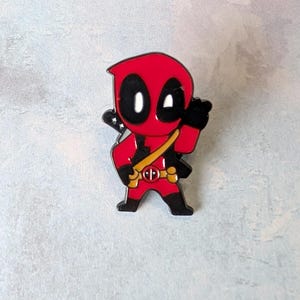 May include: A red and black enamel pin featuring a cartoon version of Deadpool. The pin has a yellow strap and a black sword on the back. The character is in a standing pose with one hand raised.