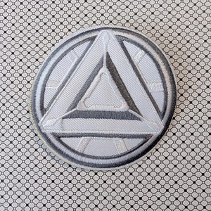 May include: A circular, embroidered patch featuring a white and gray geometric design. The design includes a central triangle and a surrounding circle, all with detailed stitching. The patch is on a patterned background.