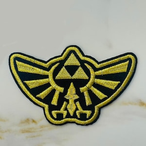 May include: A gold embroidered patch featuring the iconic Triforce symbol from the Legend of Zelda video game series. The Triforce is surrounded by a stylized winged design, with a black background.