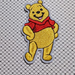 May include: Embroidered patch featuring a smiling yellow bear wearing a red shirt and a gray border. The bear is standing with its arms at its sides. The patch is on a patterned background.