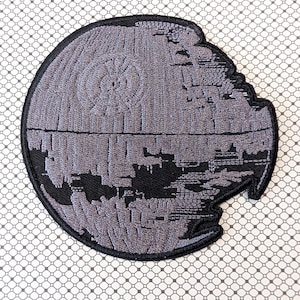 May include: Embroidered patch featuring a gray Death Star design with black accents. The patch is circular with a detailed depiction of the space station, including its surface features and a partially destroyed section. Ideal for fans of science fiction.