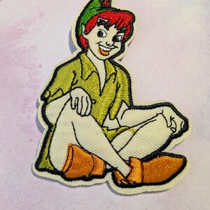 May include: Embroidered patch featuring a seated cartoon character with red hair, wearing a green hat and shirt, and orange shoes. The character is outlined in black and has a mischievous grin. The patch is on a light-colored fabric.