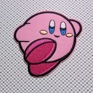 May include: Pink embroidered patch featuring a cartoon character with large blue eyes, a smiling mouth, and a protruding pink tongue. The character has a rounded body with small ears and is outlined in black. The patch is on a patterned background.