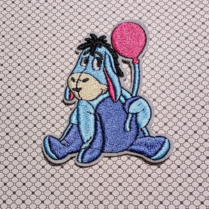 May include: Embroidered patch featuring Eeyore from Winnie the Pooh. The patch depicts Eeyore in shades of blue, with a pink balloon. The patch is outlined in a light gray color.